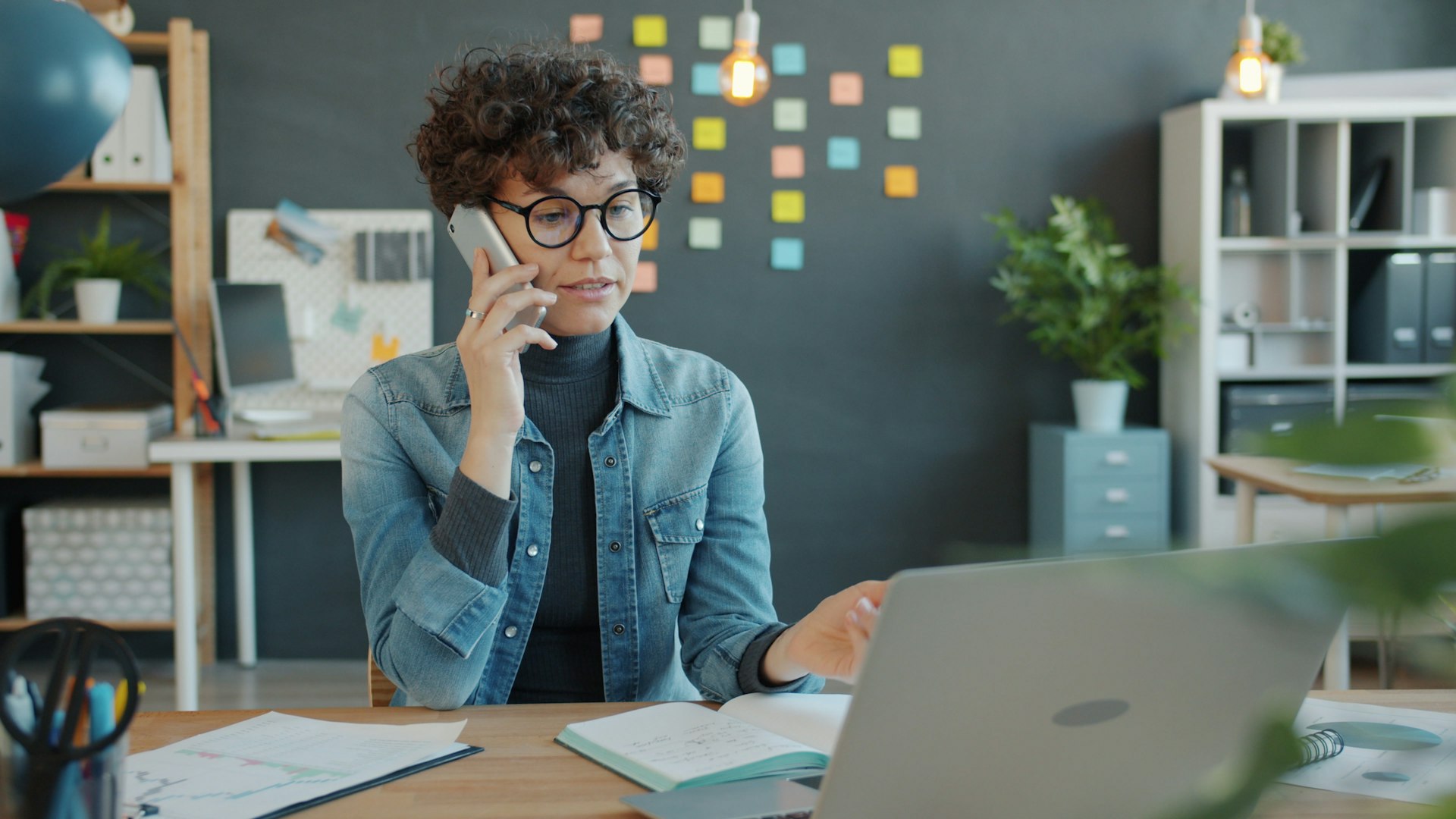 Woman with curly hair and glasses talks on phone while working on a laptop at a desk reflecting work overload. Background features colorful sticky notes on a dark wall.