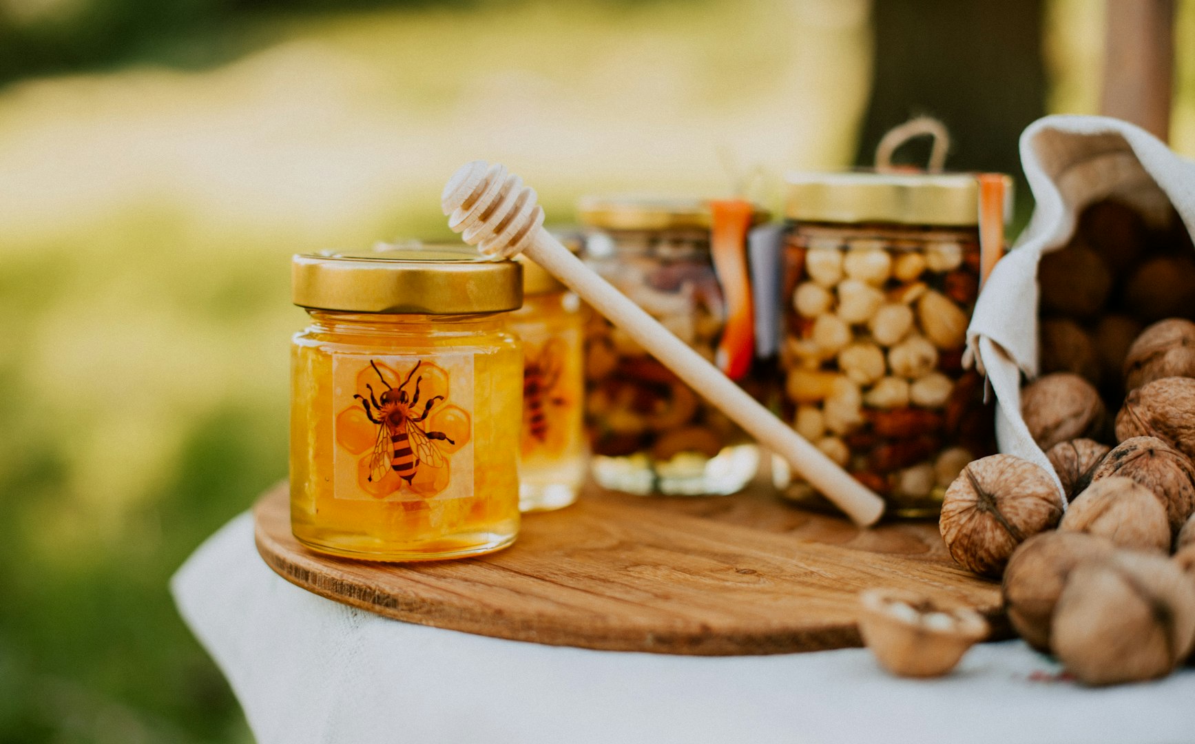 Jars of honey and nuts on a wooden board outdoors. A honey dipper leans on a jar featuring a bee design. Shelled walnuts rest nearby, conveying a rustic, natural vibe.