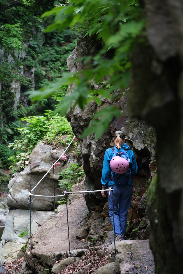 A woman in outdoor gear hikes along a narrow, rocky trail in a lush, green forest featuring slow travel activities. She carries a pink helmet, conveying a mood of adventure.