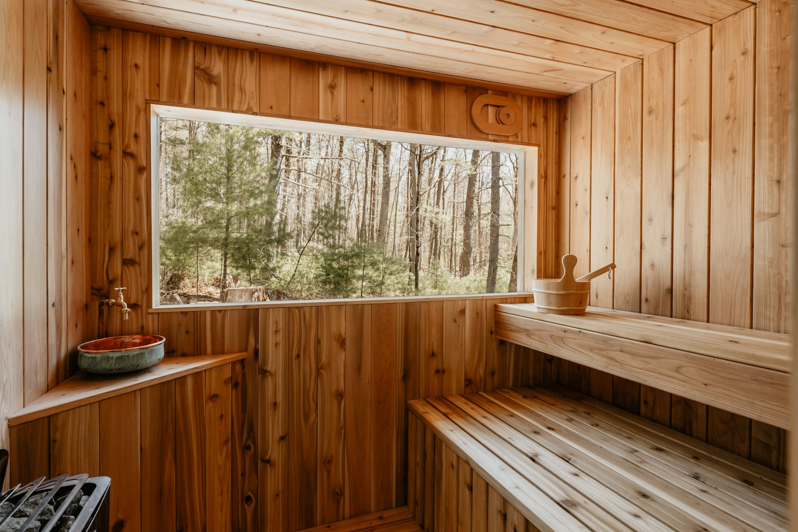 A wooden sauna interior with a large window showing a forest view. A bench and a wooden bucket add a serene, warm atmosphere, suggesting relaxation.