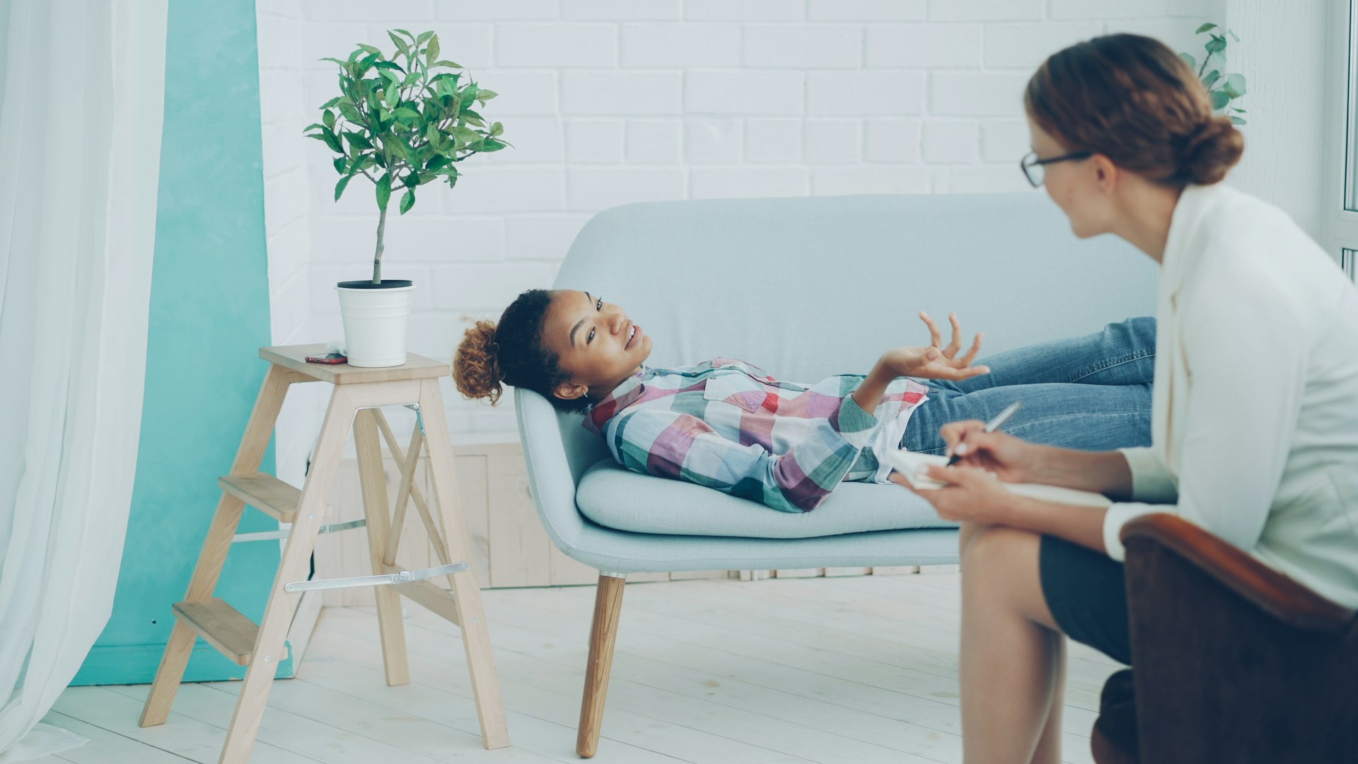 A woman in a plaid shirt lies on a sofa, gesturing while talking to a seated female therapist taking notes. The room is bright with a calm ambiance.